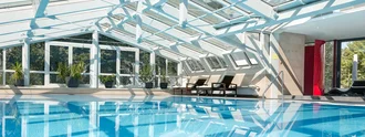 Ein großer, heller Innenpool mit klarem Wasser, umgeben von modernen Fenstern und Pflanzen. Die Architektur ist lichtdurchflutet, mit einem schrägen, transparenten Dach und gemütlichen Liegen am Rand des Pools.