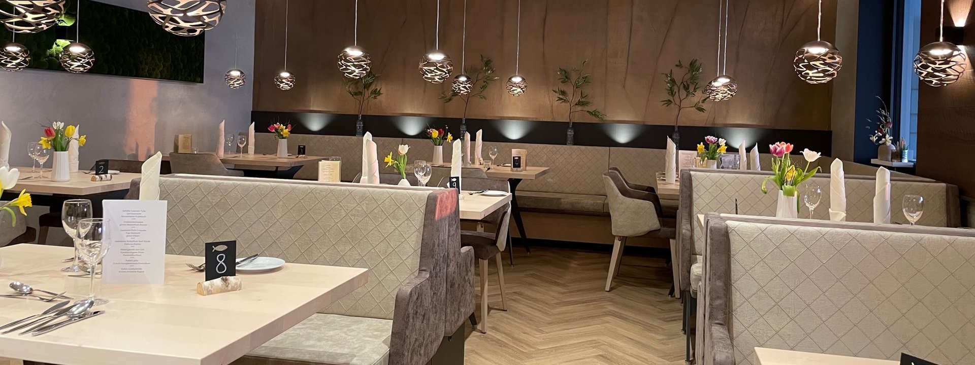 Speisesaal des Restaurants. Der Raum besteht mehreren Tischen und Stühlen in den Farben braun und beige. Silberne Lampen hängen von der Decke.